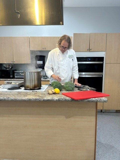Chef Chris cooking at a kitchen island with a steaming pot and fresh broccoli during a demonstration.