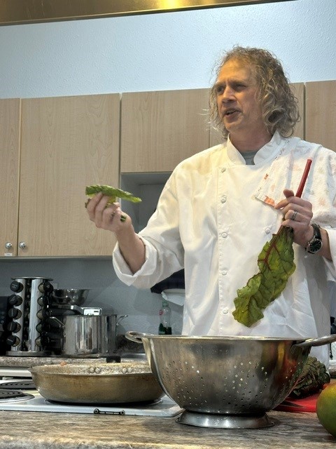 Chef Chris, in chef's whites, holds a leaf of Swiss chard while leading a hands-on cooking class in a kitchen.
