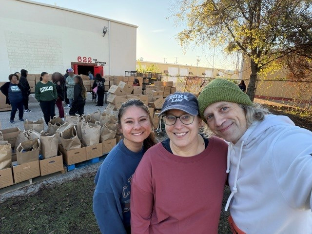 Kind Cooking representatives smiling at an outdoor community food distribution, with bagged and boxed food behind them.