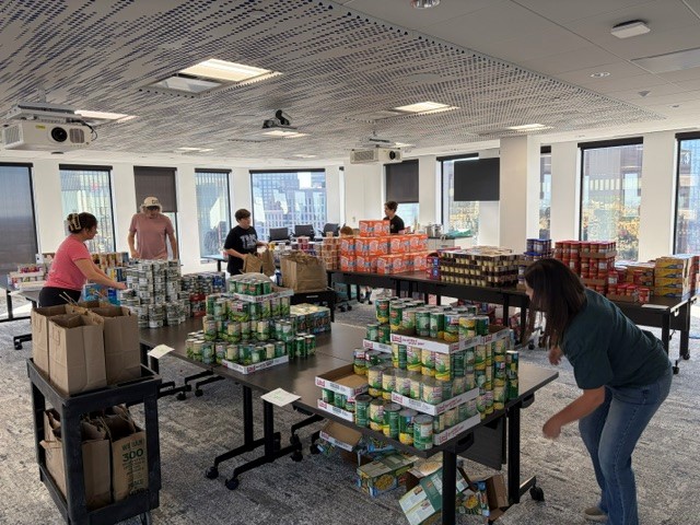 Volunteers sort donated canned goods across several tables in a large room while preparing food bags.
