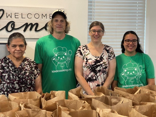 Four people, two in Kind Cooking t-shirts, standing behind packed food bags beneath a Welcome Home sign.
