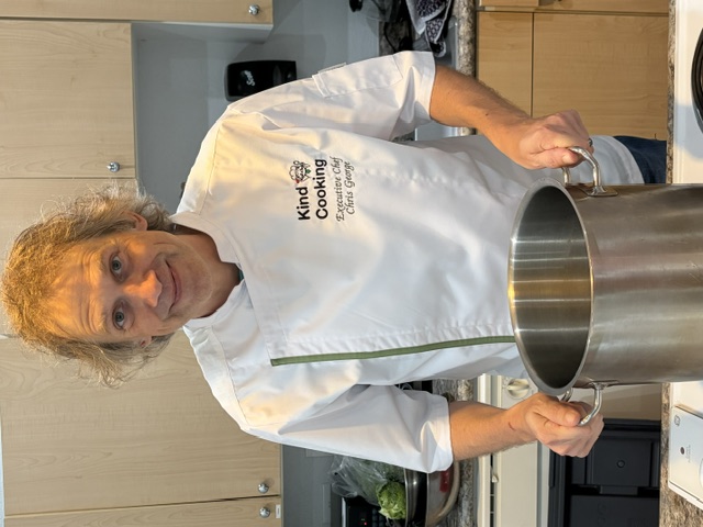 Chef Christopher George, founder of Kind Cooking, in a Kind Cooking chef's jacket behind a stockpot in a kitchen